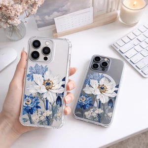 May include: Clear phone cases with floral designs. One case is held in a hand, the other is on a white surface. The floral design features white and blue flowers with green stems and leaves. The cases have a clear, protective bumper.