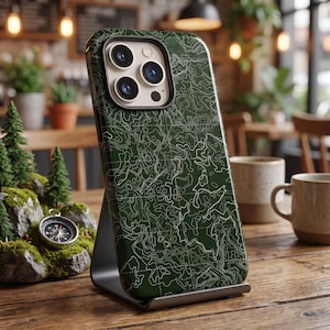 Può includere: Una cover per smartphone verde scuro con un motivo a linee topografiche bianche e una griglia. La cover presenta un ritaglio rettangolare per la fotocamera con tre lenti circolari e un flash, posizionata su un supporto metallico.