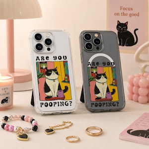 May include: Two phone cases featuring a black and white cat illustration and the text "ARE YOU POOPING?". One case is clear, the other is grey. A candle, jewellery, and a small sign with a cat graphic are also visible.