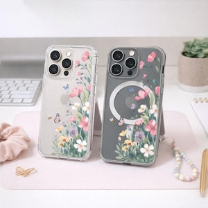 Könnte beinhalten: Zwei Handyhüllen, eine transparente und eine graue, mit Blumen- und Schmetterlingsmustern. Die transparente Hülle hat rosa Blumen, während die graue ein ähnliches Design aufweist. Beide Hüllen haben Schutzstoßstangen und sind auf einer rosa Oberfläche.