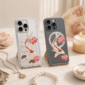 May include: Two phone cases featuring a koi carp design. One is clear, the other grey. Both cases display a white and orange koi carp, pink lotus flowers, and gold detailing. The cases are presented on gold stands.