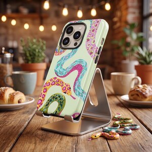 Peut inclure: Une coque de smartphone sur fond vert clair avec un motif coloré de serpents sinueux dans des tons bleu, rose, jaune et vert. La coque est fixée sur un support en métal argenté, révélant une découpe pour triple objectif photo au dos.