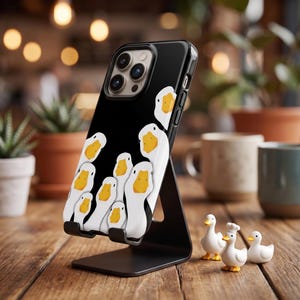 Puede incluir: Una funda de smartphone negra con un patrón repetitivo de cabezas de pato blancas de dibujos animados con picos amarillos brillantes. La funda está montada en un soporte de metal negro, mostrando la lente de la cámara y el diseño gráfico.