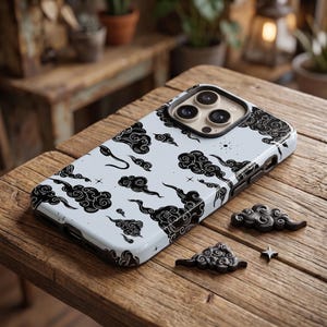 以下が含まれることがあります： 水色のスマートフォンケースで、黒いインクで描かれたような様式化された雲と小さな四芒星の繰り返し模様が特徴です。ケースにはカメラ用の隆起した切り抜きがあり、2つの小さな黒い雲の形をした装飾品が添えられています。