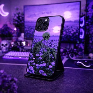 以下が含まれることがあります： 紫色の花畑と夕焼けを背景に、骸骨が座っているデザインの黒い携帯電話ケース。 ケースは黒いスタンドに立てかけられており、背景には紫色のコンピューターモニターとキーボードがあります。