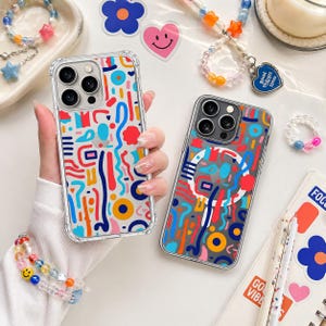 Könnte beinhalten: Zwei transparente Handyhüllen mit farbenfrohen abstrakten Designs. Eine Hülle zeigt rote, blaue, gelbe und orangefarbene Formen auf weißem Hintergrund. Die andere Hülle hat ein ähnliches Design auf grauem Hintergrund. Armbänder und Aufkleber sind ebenfalls zu sehen.
