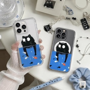 Puede incluir: Dos fundas transparentes para teléfono con un diseño de gato negro. El gato está representado en una escena de agua azul con peces pequeños. Una funda la sostiene una persona. Otros accesorios incluyen un bolígrafo, una goma para el pelo y una pulsera de cuentas.
