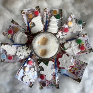 Pode incluir: Uma coleção de oito kits de artesanato dispostos em círculo em torno de uma caneca de vidro com café. Cada kit contém uma figura de Natal em cerâmica branca, um pequeno pincel e vários potes de tinta, todos com o texto kreativ zwerge.