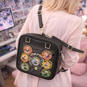May include: Black square crossbody bag with a clear front panel displaying colorful character pins and a decorative ribbon. The bag has gold-colored hardware and a black adjustable strap.