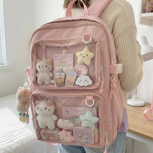 Puede incluir: Mochila rosa claro con bolsillos frontales transparentes, que muestra colgantes de gato y estrella de peluche, junto con accesorios de té de burbujas y nube. Cuenta con correas ajustables y asa superior.