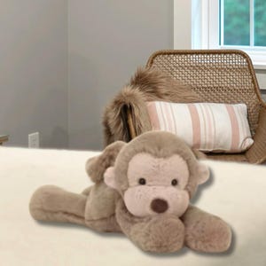 Pode incluir: Um peluche de macaco com pelo macio castanho-claro, um rosto bege mais claro e um nariz castanho-escuro. O animal de peluche está representado numa posição deitado com as patas dianteiras estendidas, orelhas pequenas e olhos escuros.
