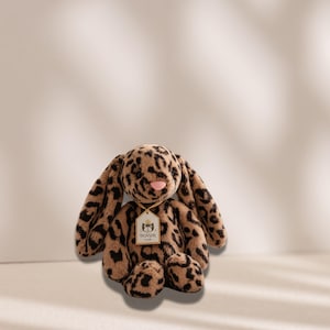 May include: A plush stuffed animal in the shape of a rabbit with long ears, covered in a brown and black leopard print pattern. The rabbit has a small pink nose and wears a beige ribbon around its neck with a white tag featuring the text JELLYCAT.