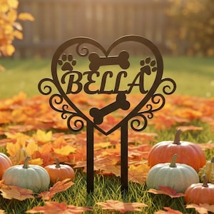 May include: A black metal garden stake with a heart-shaped design, featuring the name "BELLA", paw prints, and a bone. The stake is placed in a yard with pumpkins and autumn leaves.