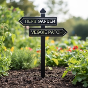Puede incluir: Señal de jardín de metal negro con flechas direccionales. El letrero superior dice "HERB GARDEN" y el letrero inferior dice "VEGGIE PATCH". La señal se coloca en un parterre con varias plantas y flores verdes.
