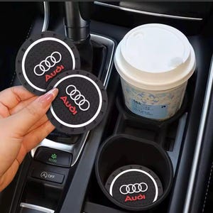 May include: Black car coasters featuring the Audi logo in white and red. The coasters are circular and designed to fit car cup holders. A white paper cup with a lid is in a cup holder. The car's interior is black and grey.