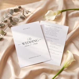 May include: A white wedding program card featuring black text that reads Welcome to The Wedding Sep 11 2026 Aurora & Phillip Washington Ave, NY, with an order of ceremony and wedding party list on a second card, accented by white calla lilies.