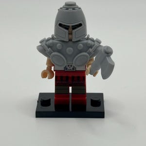 May include: A grey Lego knight figurine, featuring a helmet, shoulder armour, and a weapon. The knight has a red and brown torso and legs, and stands on a black base. The figurine is set against a white backdrop.