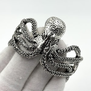 Peut inclure: Un bracelet manchette en métal argenté arborant un motif détaillé de pieuvre. La pieuvre présente une tête texturée et de multiples tentacules incurvés qui s'enroulent autour de la bande, révélant des motifs de ventouses sur toute la surface.