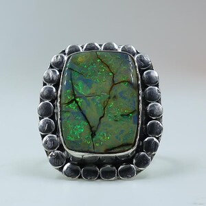 May include: A silver ring featuring a rectangular, dark green, iridescent opal stone with visible cracks and flashes of bright green and blue light. The stone is set in a bezel surrounded by a decorative border of small, rounded silver spheres.