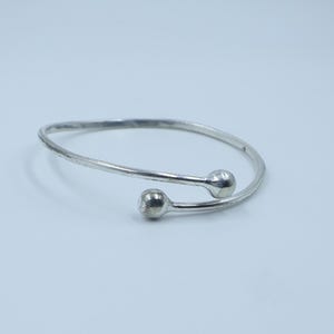 May include: A silver-colored open cuff bracelet with two ball-shaped ends. The bracelet has a simple, elegant design and is set against a plain background. The metal appears to be slightly textured.