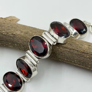 May include: A silver bracelet featuring oval-cut, deep red gemstones. The gemstones are set in individual silver bezels and linked together. The bracelet is displayed on a piece of wood against a white background, highlighting the jewelry's design and color.