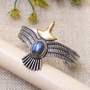 May include: A silver-toned cuff bracelet featuring an eagle design with spread wings. The eagle has a polished gold-toned head and chest, with an oval-shaped, iridescent blue labradorite gemstone set in the center of its body. The wings are textured.