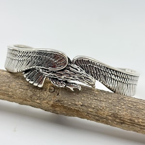 Puede incluir: Una pulsera de puño plateada con un diseño de águila. La pulsera presenta alas detalladas y una cabeza de águila esculpida. Las alas se envuelven alrededor de la muñeca, creando un accesorio llamativo. La pulsera se muestra sobre un trozo de madera.