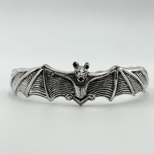 Op de afbeelding: Een zilverkleurige armband met een gedetailleerd vleermuisontwerp met uitgespreide vleugels. Het lichaam van de vleermuis is gecentreerd op de armband, met ingewikkelde details op de vleugels en het lichaam. De armband heeft een gebogen vorm, ontworpen om de pols te passen.