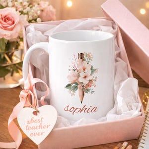 Puede incluir: Taza de cerámica blanca con un diseño floral y de lápiz, personalizada con el nombre "Sophia". La taza se presenta en una caja de regalo rosa con una etiqueta en forma de corazón que dice "best teacher ever".