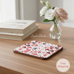 May include: Square coaster with a heart pattern in red, black, and pink on a white background. The coaster has a brown border and is on a wooden surface. A vase of flowers and a stack of books are in the background.
