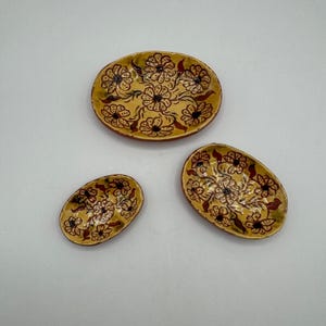 May include: Three handmade ceramic dishes. The dishes are oval-shaped with a yellow base and a floral design in brown and black. The largest dish is at the top, with two smaller dishes below.