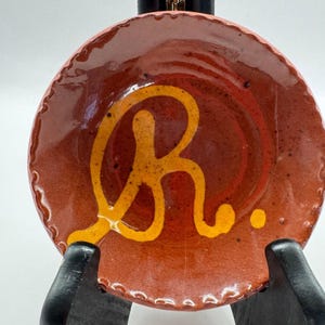 May include: A small, round, handmade ceramic dish with a reddish-brown glaze. The dish features a large, stylized yellow-orange letter "R" in the center. The rim has a scalloped edge. The dish is displayed on a black stand.