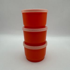 Puede incluir: Tres recipientes apilados de color naranja con tapas de color rosa claro. Los recipientes son cilíndricos con un borde ligeramente ensanchado. Las tapas encajan perfectamente. Fabricados con material opaco.