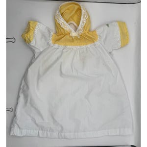 Könnte beinhalten: Ein weißes Kleid mit kurzen Ärmeln, das gelb-weiß karierten Stoff an der Kapuze und den Ärmelbündchen aufweist. Das Kleidungsstück ist mit weißer Spitzenborte an der Kapuze, der Passe und den Ärmelkanten verziert und hat eine kleine gelbe Schleife.