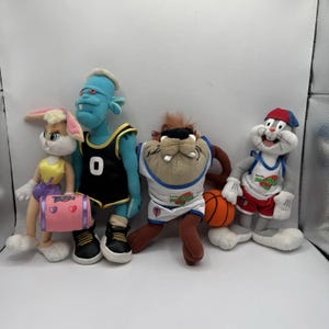 May include: A selection of soft toys depicting characters from the film Space Jam. The toys include Lola Bunny, a blue monster, Taz, and Bugs Bunny. The characters are wearing basketball kits and holding a basketball.