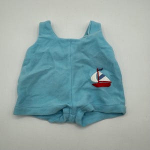 Könnte beinhalten: Hellblaue Baby-Spielhose mit Segelboot-Applikation. Die Spielhose hat ein ärmelloses Design und einen runden Ausschnitt. Das Segelboot ist rot, weiß und blau. Die Spielhose ist aus weichem Frotteematerial.