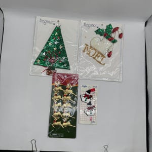 May include: Four packages of Christmas-themed fabric appliques. One features a green sequined tree, another a white stocking with the word NOEL and a candy cane, a third contains ten gold reindeer, and the fourth shows two small snowman figures.