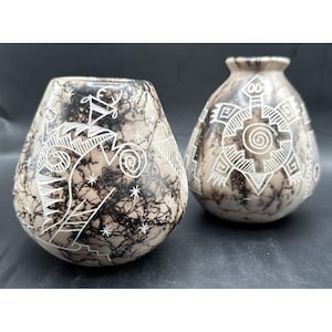 May include: Two decorative, hand-carved stone vases. The vases have a teardrop shape and are beige with black veining. One vase features white geometric designs, while the other has a spiral and star pattern. The vases are displayed on a dark surface.
