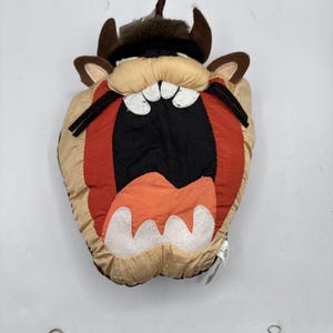Könnte beinhalten: Ein Plüschkissen in Form des Kopfes des Tasmanian Devil. Das Kissen hat ein beiges Gesicht, braune Hörner und einen weit geöffneten roten Mund mit weißen Zähnen. Schwarze Akzente umranden die Augen und den Mund.