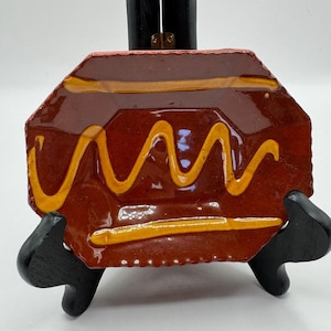 May include: A handmade, ceramic plate with a unique, geometric shape. The plate is glazed in a rich, reddish-brown color, with a wavy, mustard-yellow design. The plate is displayed on a black stand.