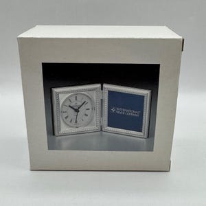 Può includere: Una scatola di cartone bianco con l'immagine stampata di una cornice doppia incernierata color argento. Un lato contiene un orologio analogico rotondo con numeri romani, mentre l'altro mostra un quadrato blu con il testo International Silver Company.