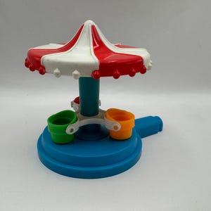 May include: A plastic toy carousel featuring a red and white striped canopy top, a central teal pillar, and a blue circular base. The ride includes small, colorful buckets in green, orange, and red attached to the rotating center mechanism.