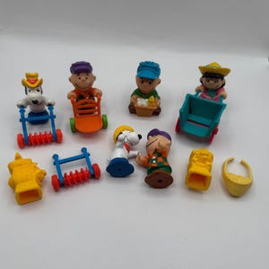 May include: A collection of colorful Peanuts character figurines. The figurines include Snoopy, Charlie Brown, and other characters, some with accessories like wagons and a fire hydrant. The figures are made of plastic and are on a white background.