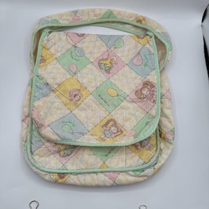 Peut inclure: Un porte-bébé matelassé avec un motif patchwork pastel. Le porte-bébé présente un motif répété d'enfants de dessins animés, de ballons et le texte "Cabbage Patch Kids". Les bords sont bordés d'un tissu vert clair.