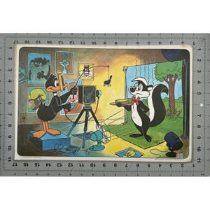 May include: A vintage cartoon print featuring Daffy Duck operating a camera on a tripod, with Pepe Le Pew holding a tray. The scene is set in a photography studio, with other Looney Tunes characters' portraits on the wall. The print has a retro aesthetic.