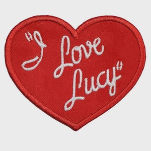 May include: A red heart-shaped patch with the phrase "I Love Lucy" embroidered in white cursive script. The patch has a stitched border and is likely designed to be sewn onto clothing or accessories.