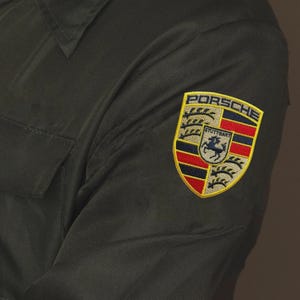 May include: Close-up of a dark green shirt with a Porsche emblem patch on the sleeve. The patch features the Porsche logo with the words "PORSCHE" and "STUTTGART" and a horse.