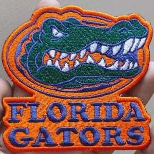 May include: Embroidered patch featuring the Florida Gators logo. The patch has an orange background with a blue and green alligator design. The words "FLORIDA GATORS" are embroidered in blue.