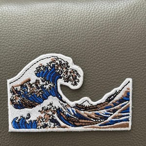 May include: Embroidered patch featuring a stylized depiction of a large ocean wave, with blue, brown, and white colors. The design includes a mountain in the background. The patch has a white border and is likely for clothing or accessories.