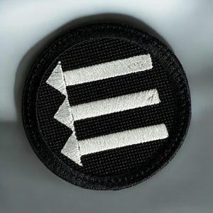 May include: Round black patch with three horizontal white lines and three white arrowheads pointing left. The patch has a black border and is likely an embroidered emblem or patch.